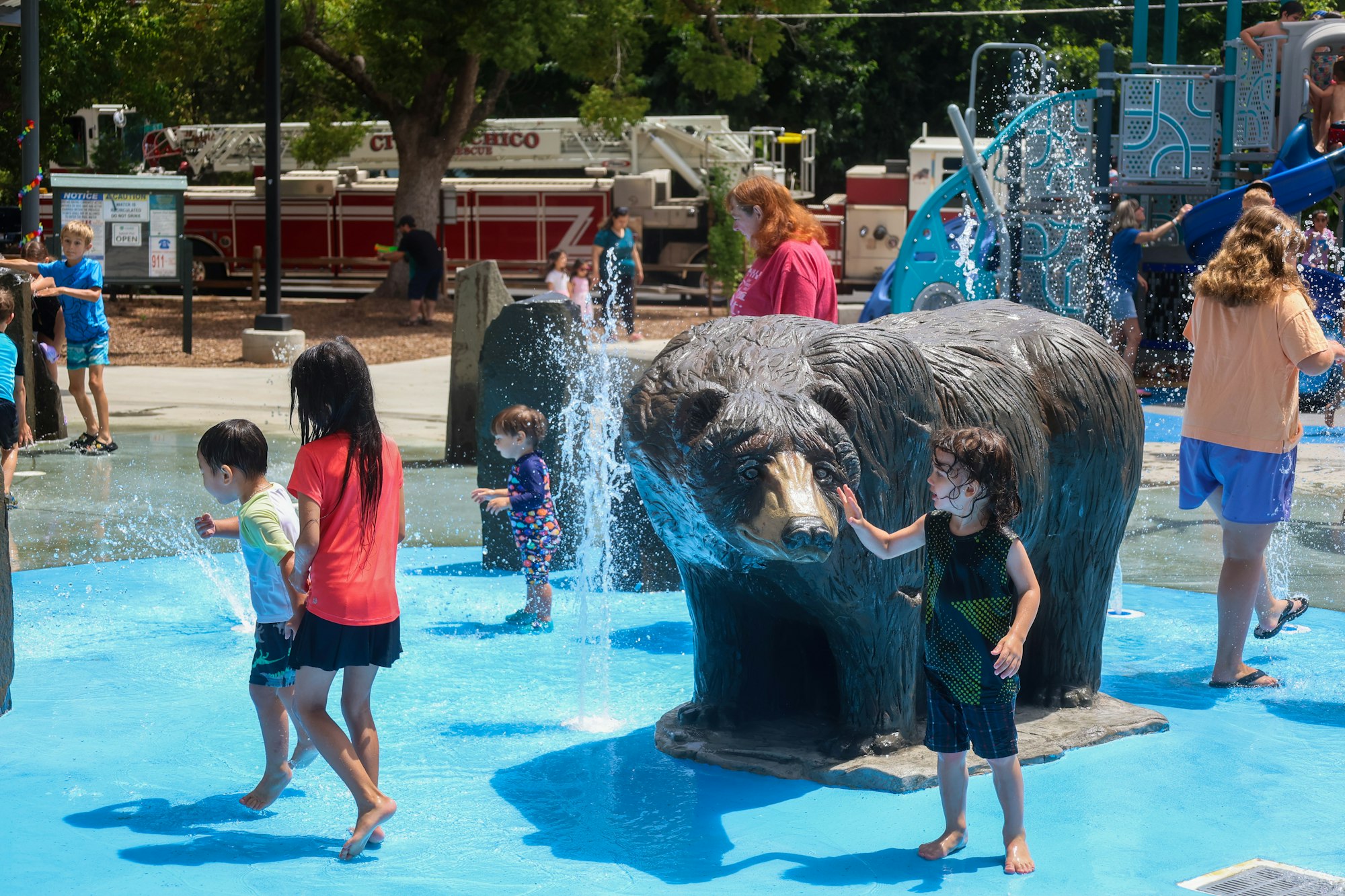 Kids playing in a water splash pad featuring a bear statue, with a playground and a fire truck in the background.