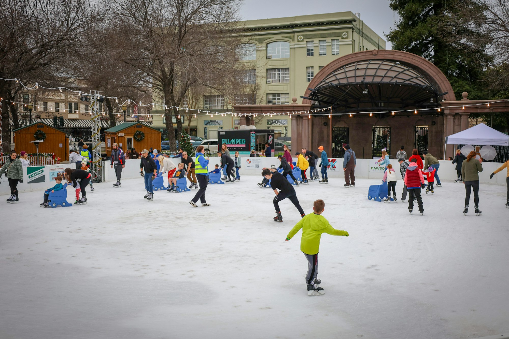 People ice skating and using aid devices on an outdoor rink, surrounded by festive decorations and trees.