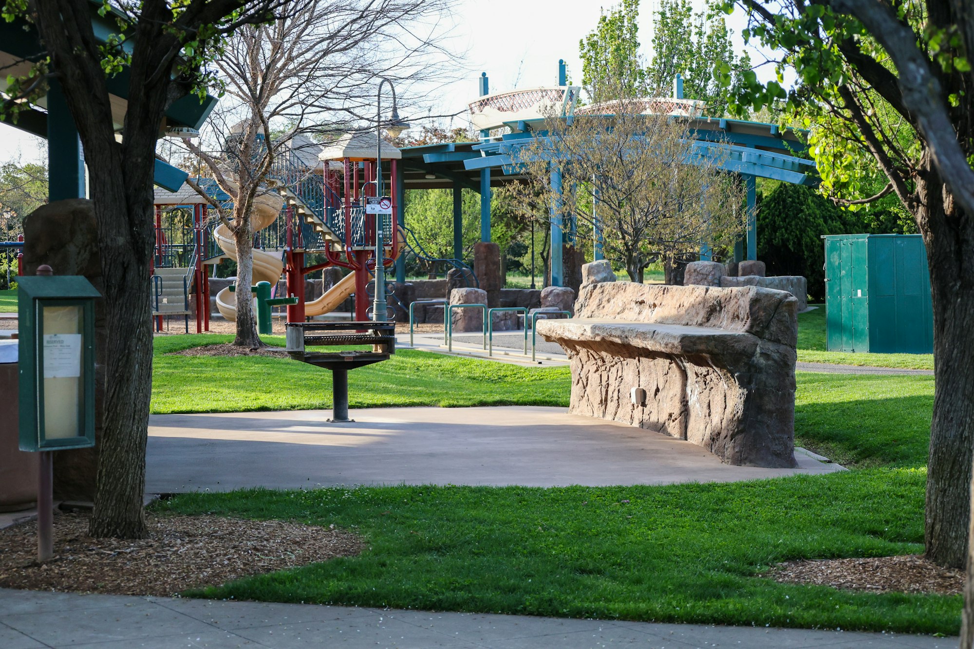 A playground with slides and climbing frames, trees, a bench, and walking paths in a park.