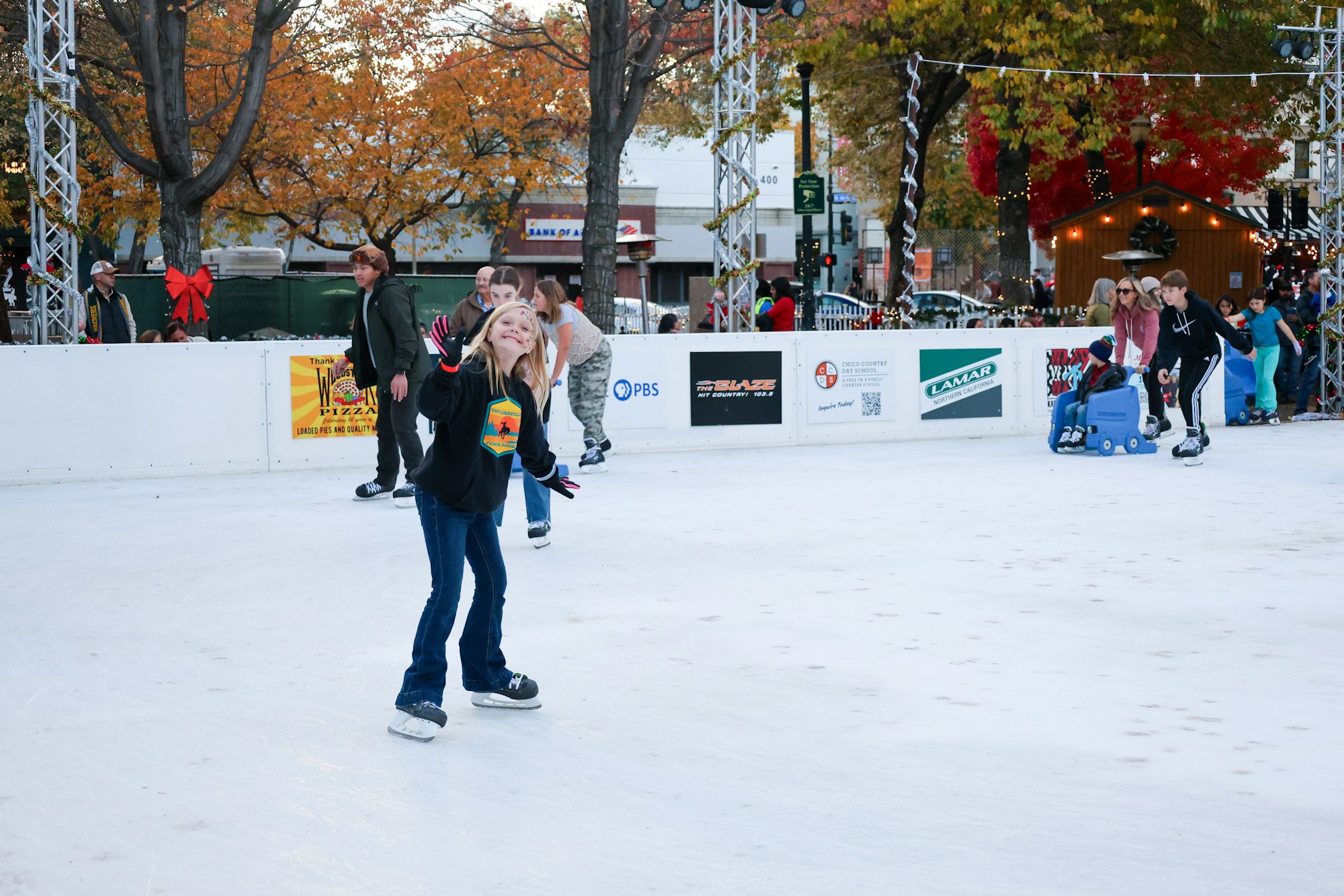 People ice skating outdoors, autumn trees, festive atmosphere.
