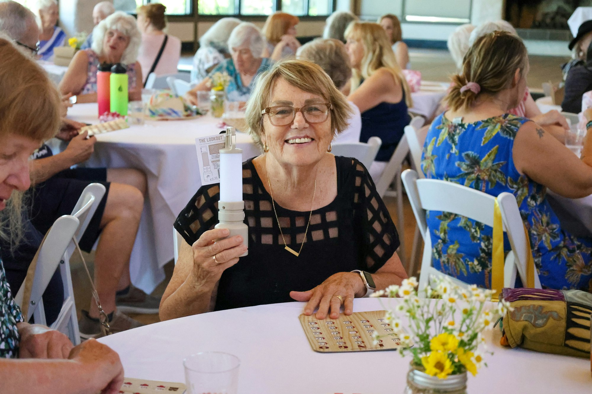 A woman smiling at a table, holding a small lamp, surrounded by people in a social gathering.