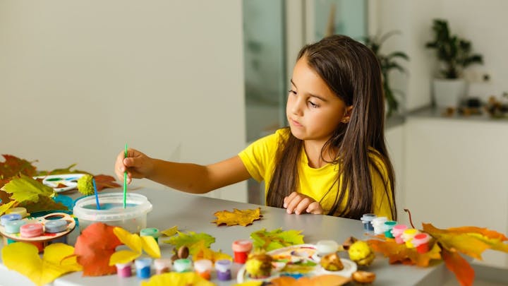A girl painting colorful leaves among art supplies on a table.