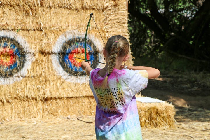 A girl aiming a bow and arrow at a target on hay bales.