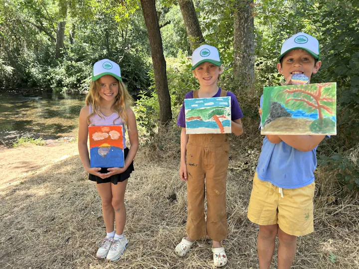 Three children hold their colorful paintings outdoors by a creek, surrounded by trees on a sunny day. They wear matching hats.