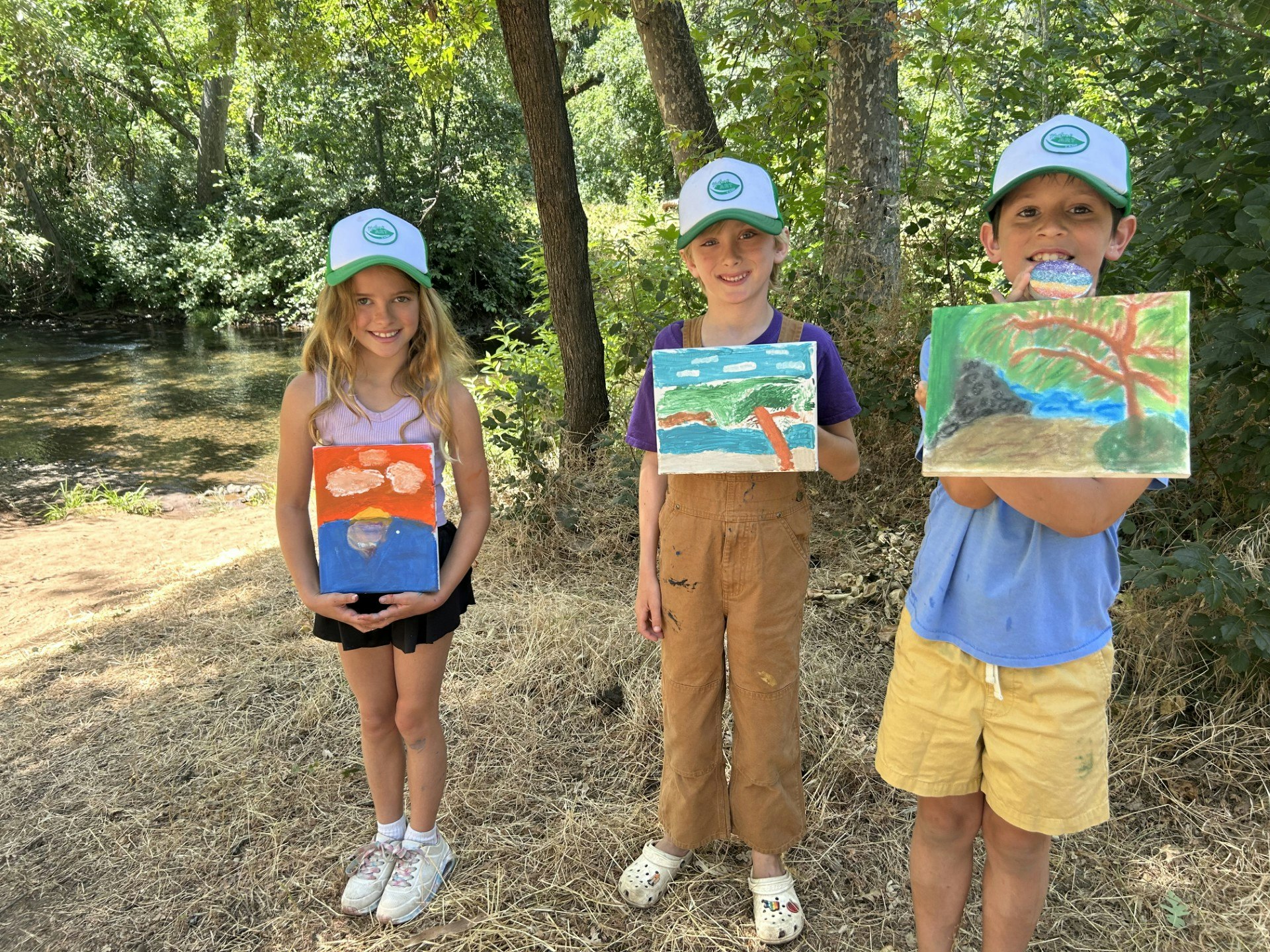 Three children hold their colorful paintings outdoors by a creek, surrounded by trees on a sunny day. They wear matching hats.
