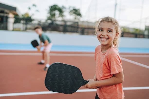 A girl smiles while holding a paddle on a court, with a boy in the background preparing to play.