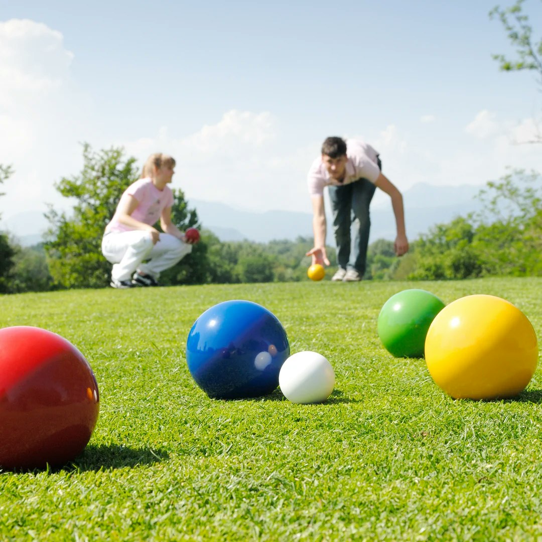 Two people are playing a lawn game on grass, with colorful balls scattered around them under a clear sky.