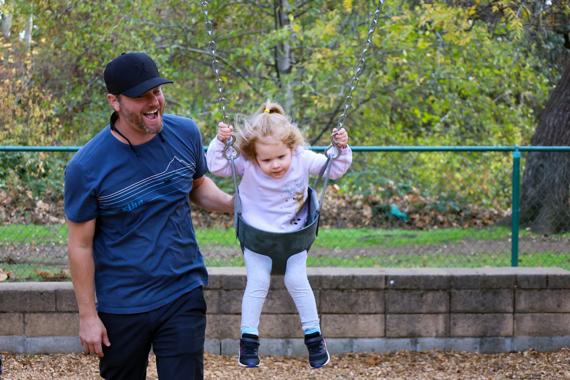 A man pushing a child on a swing, both looking happy at a park.