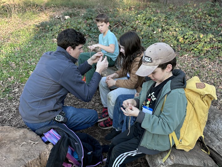 A group of kids and an adult are sitting outdoors, engaging in a hands-on activity, possibly learning a craft or skill.
