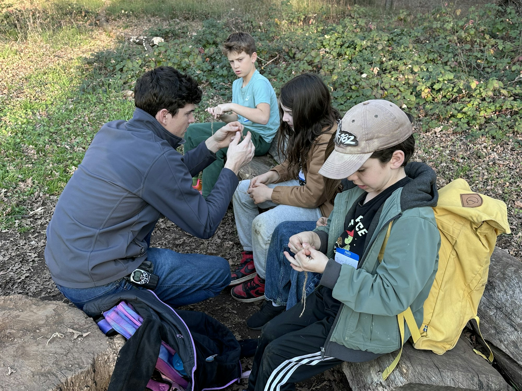 A group of kids and an adult are sitting outdoors, engaging in a hands-on activity, possibly learning a craft or skill.