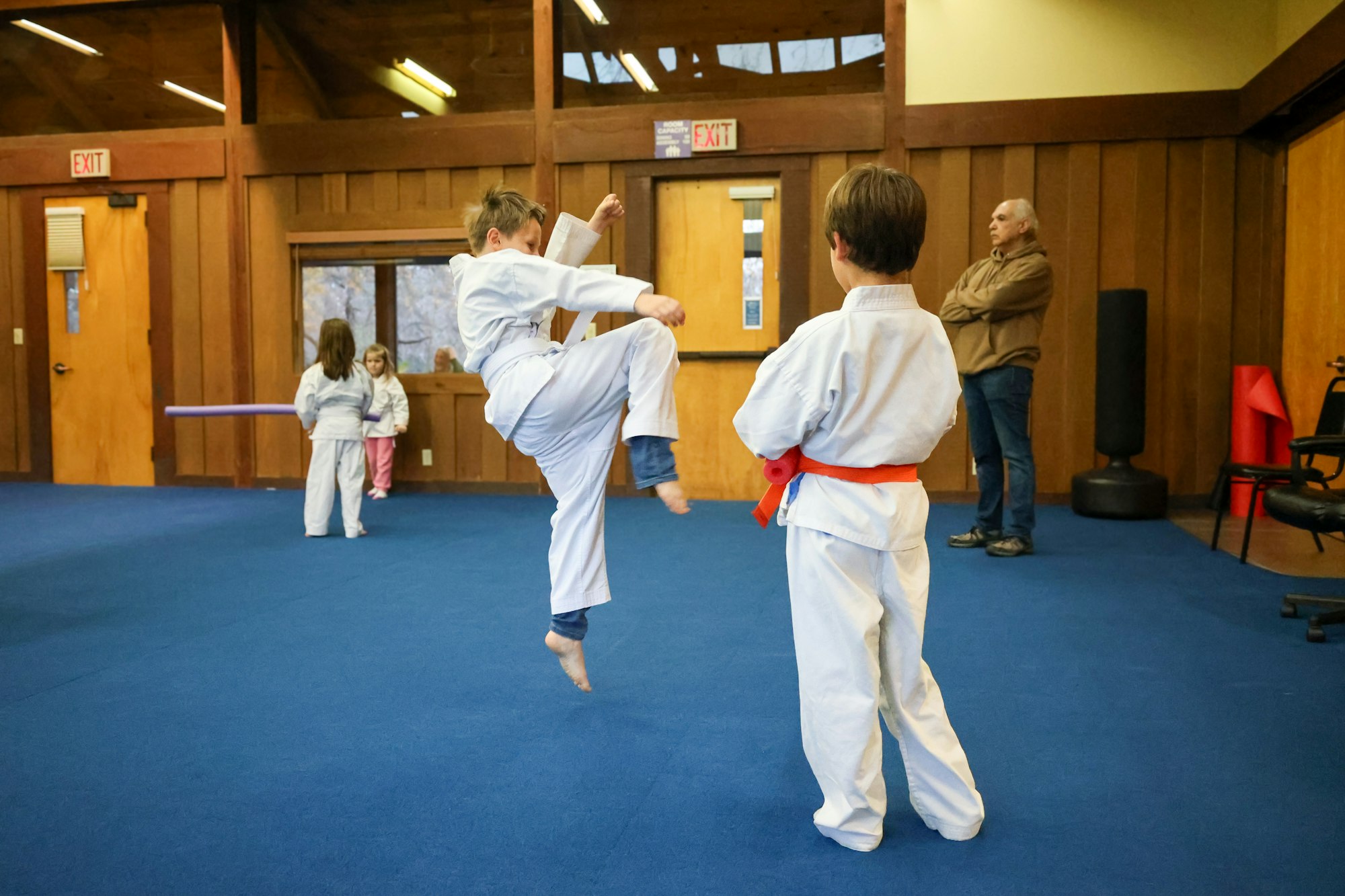 Children in karate class, one kicking high, instructor watching.