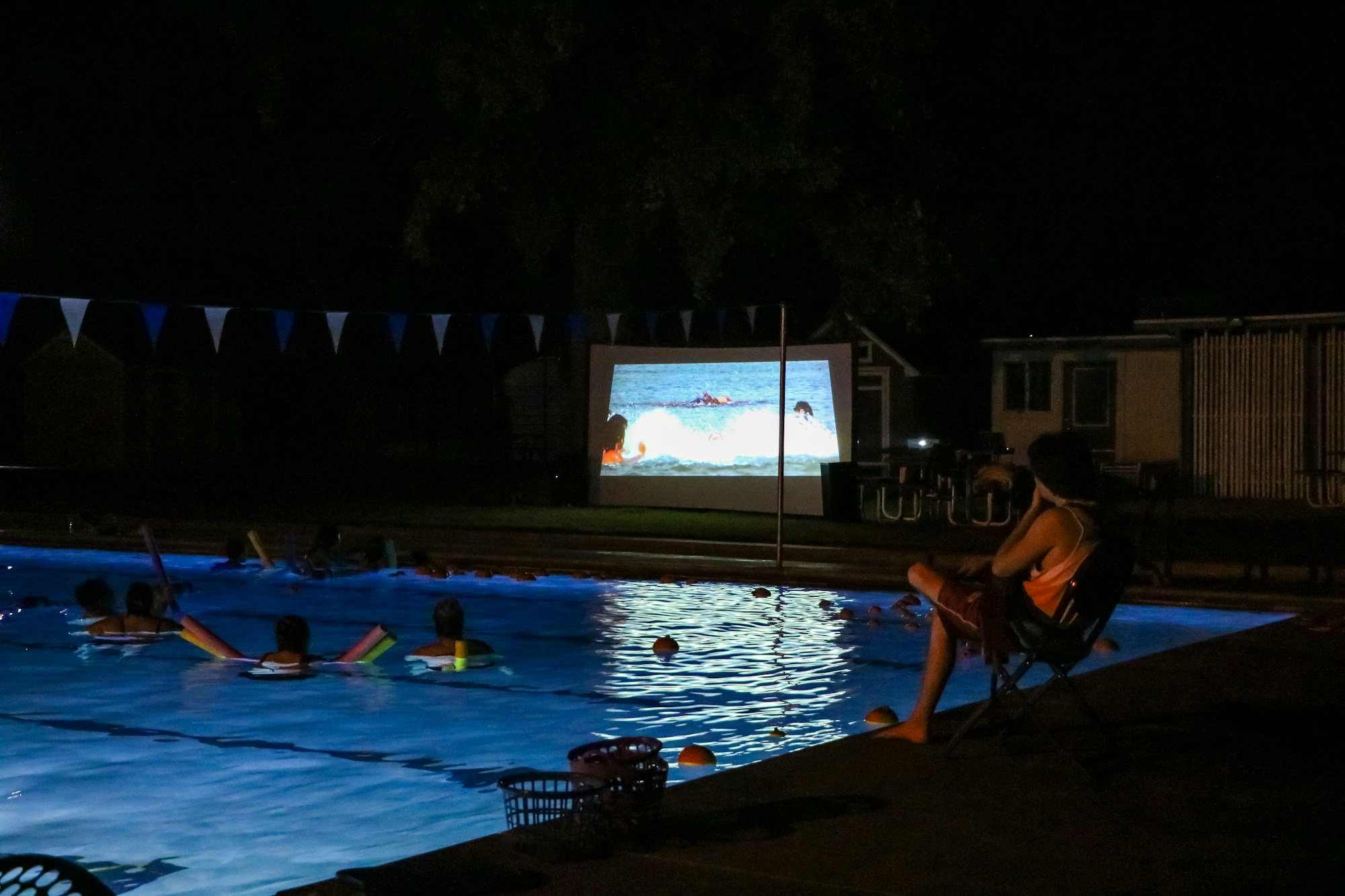 People watching a movie projected on a screen by a pool at night.
