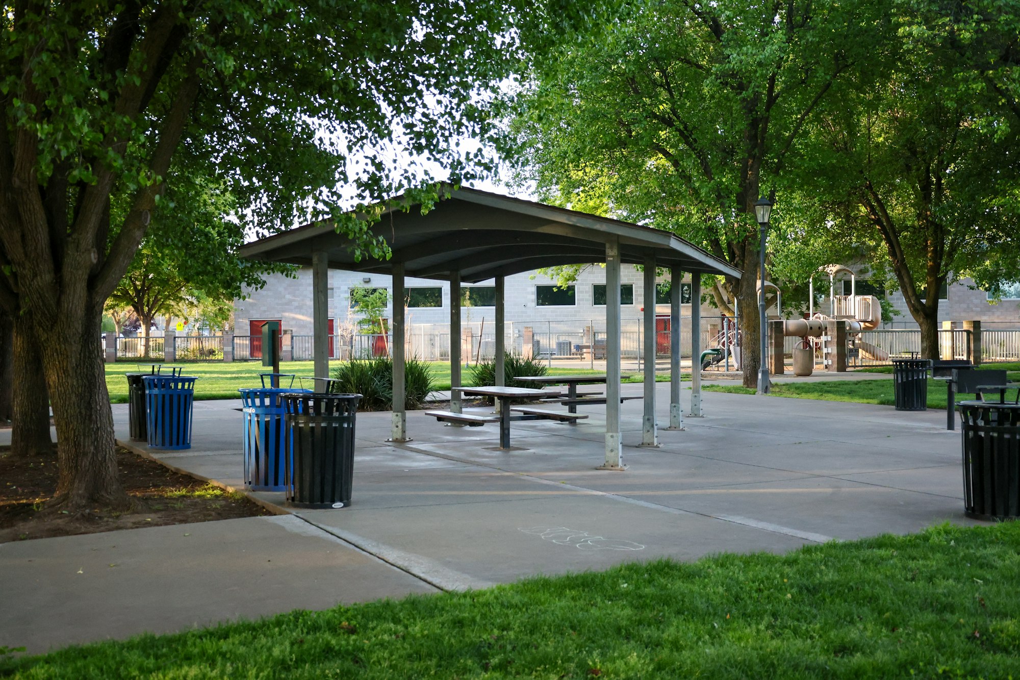 A park with picnic tables under a shelter, trash bins, trees, and a playground in the background.