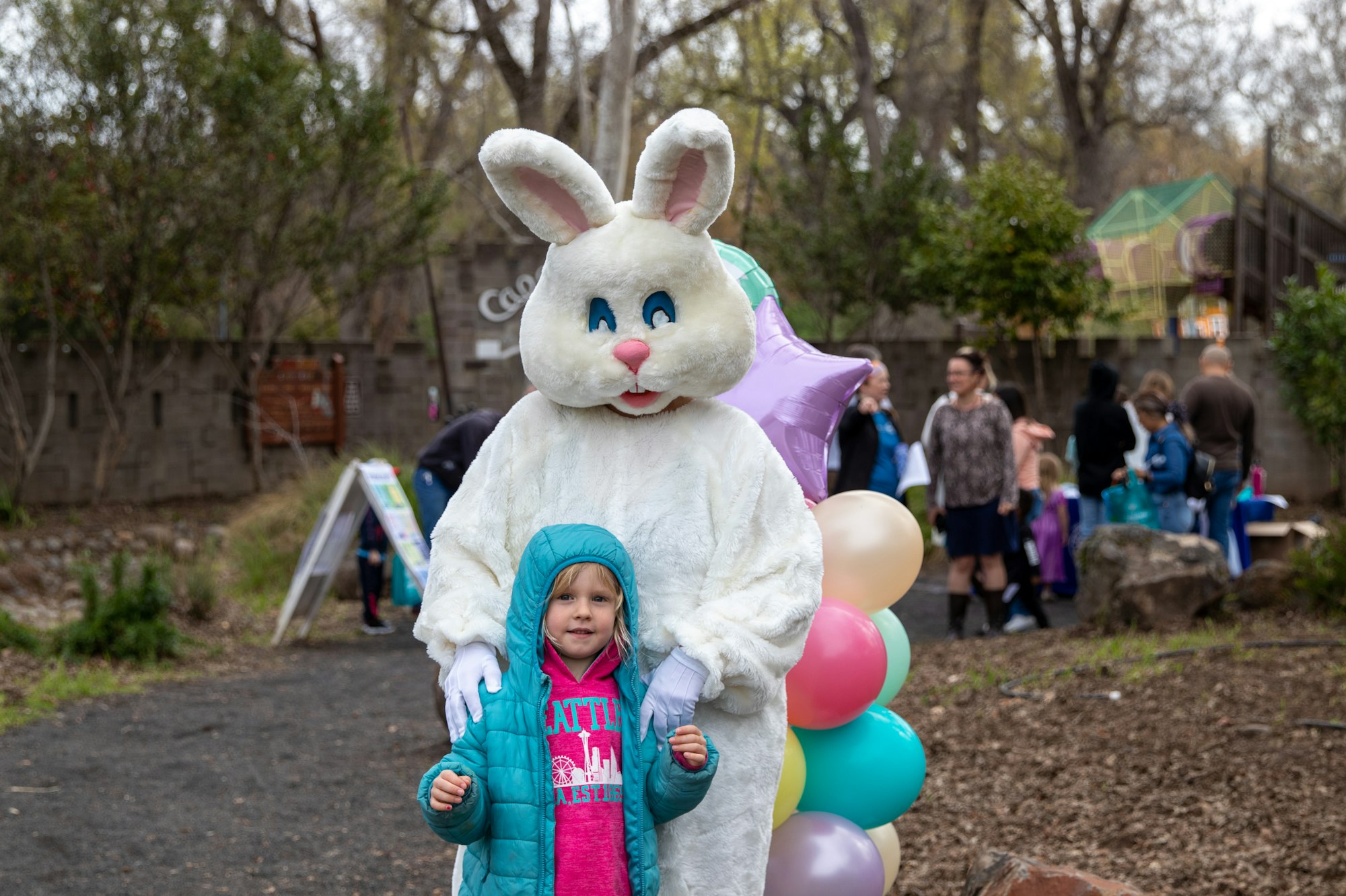 Person in bunny costume with a child in a jacket and balloons, outdoors with people in background.