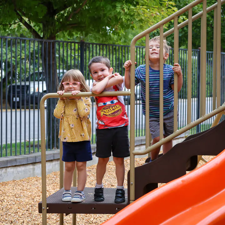 Three children standing on a playground platform, smiling and posing near a slide.