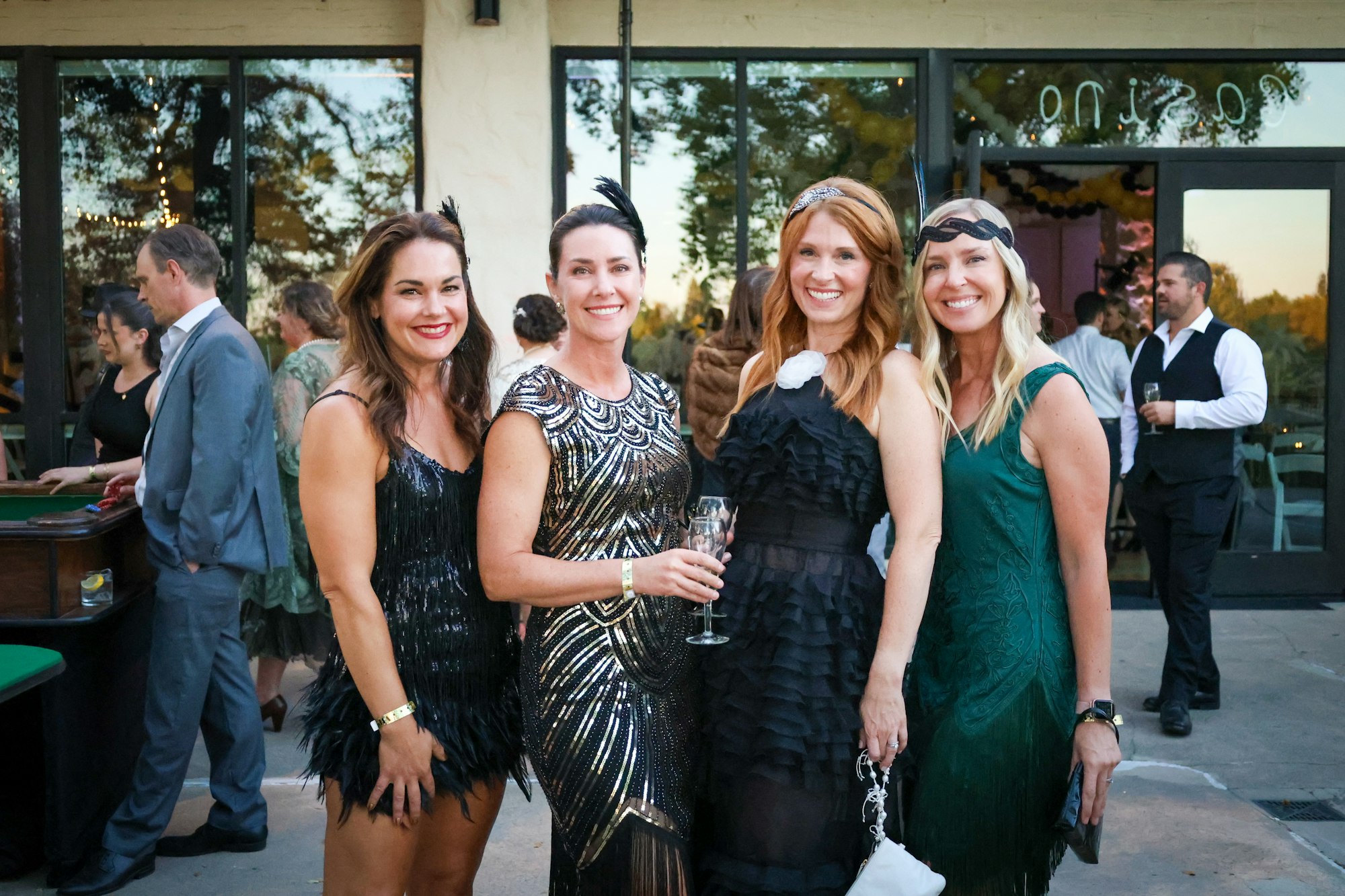 Four women in elegant dresses posing at an outdoor event with other guests in the background.