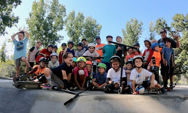 A group of kids with helmets and scooters posing together at a skate park, surrounded by trees on a sunny day.