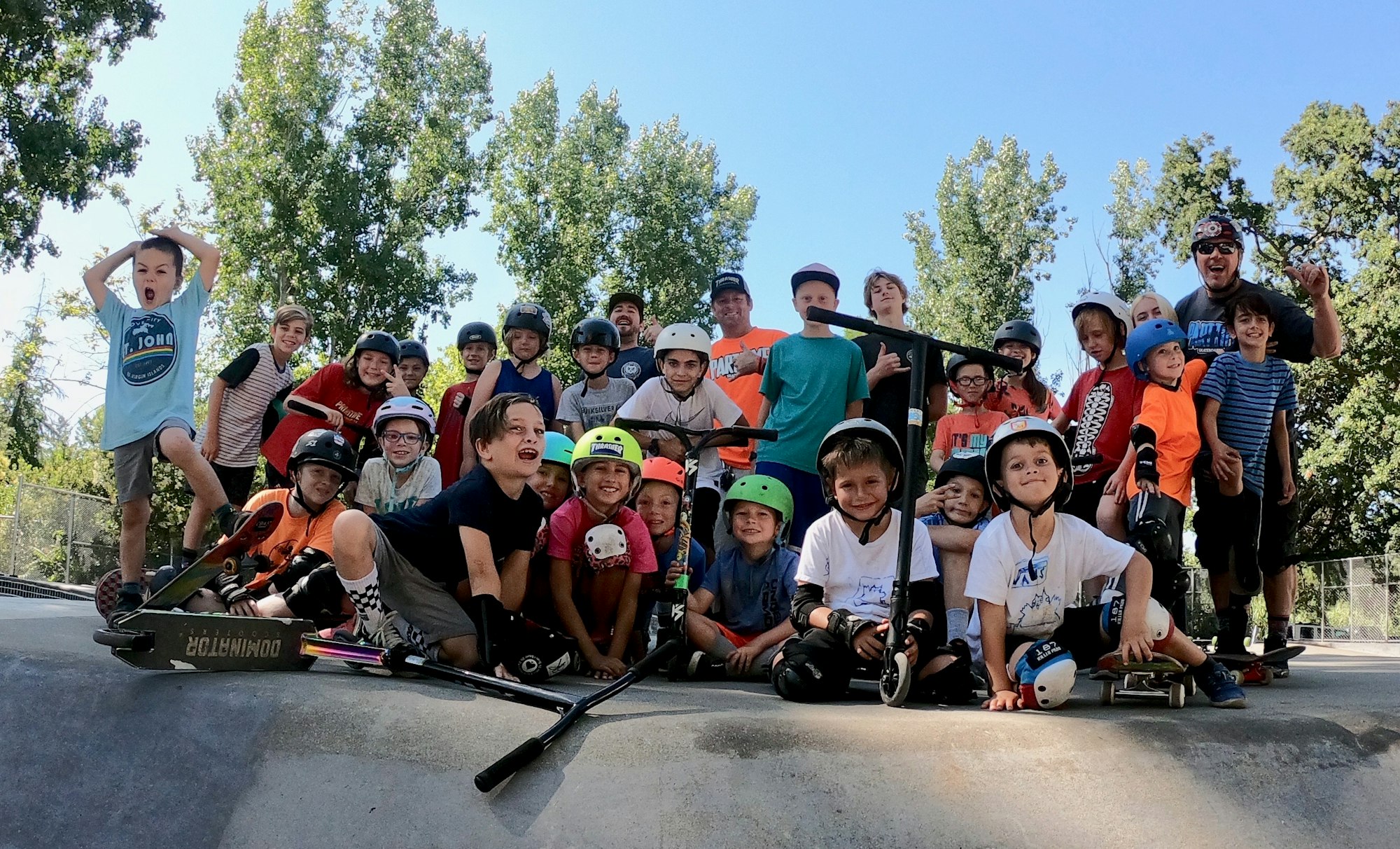 A group of kids with helmets and scooters posing together at a skate park, surrounded by trees on a sunny day.