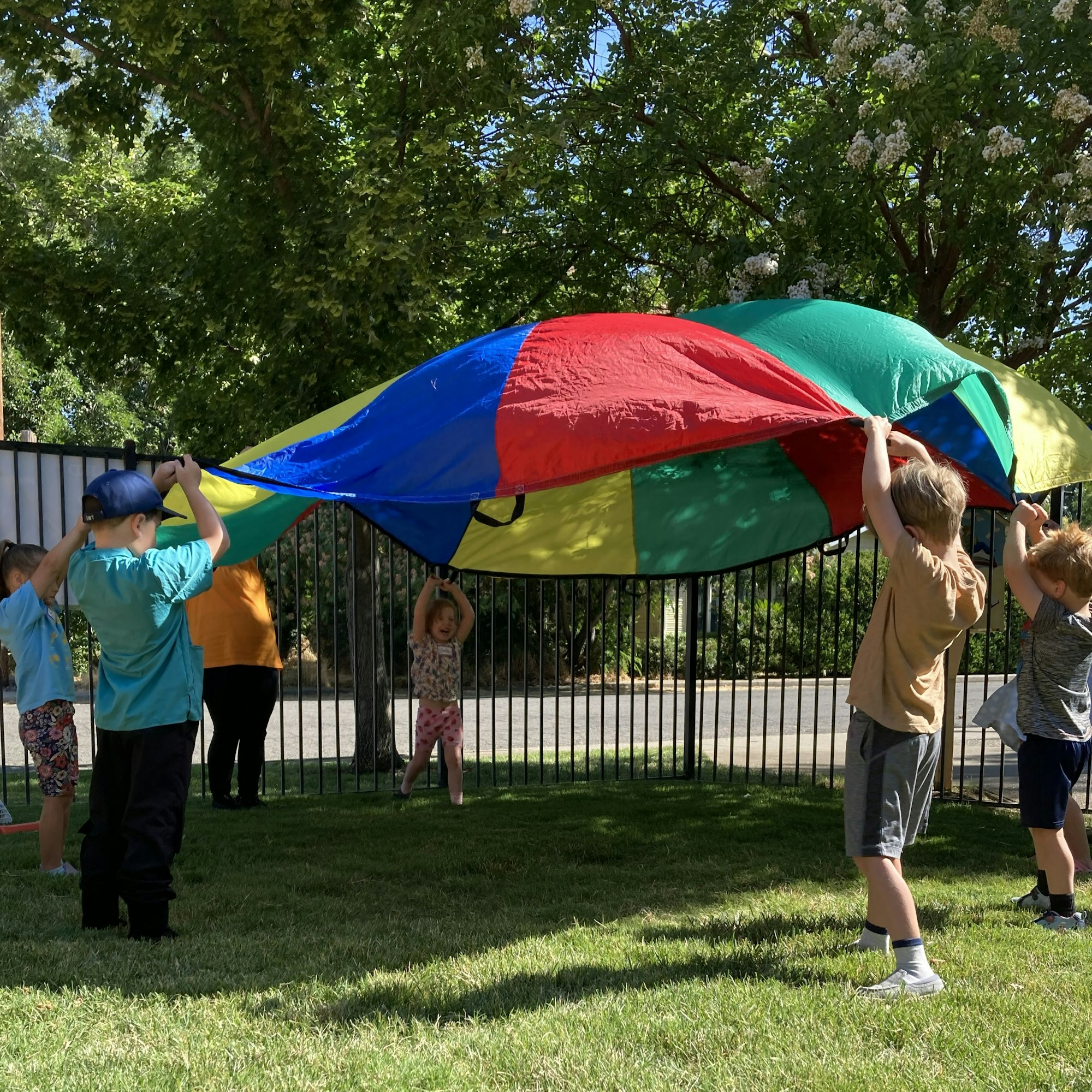 Children are playing with a colorful parachute outside, creating a fun moment under the vibrant canopy.
