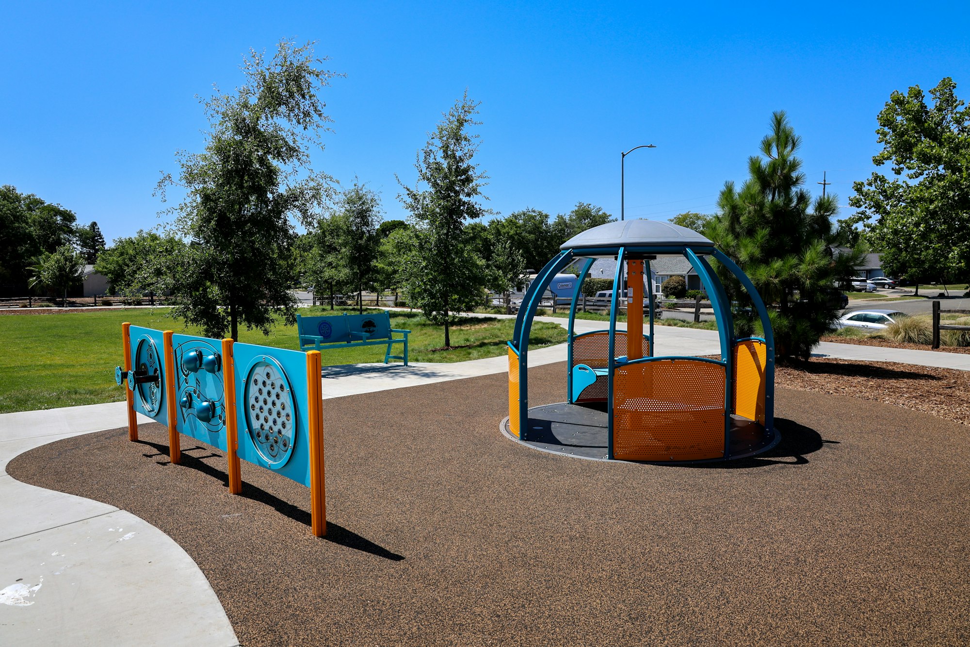 A playground with interactive panels, a round play structure, bench, and trees under a clear blue sky.