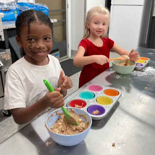 Two children are happily baking in a kitchen, mixing batter and using colorful cupcake liners. Flour is scattered on the table.