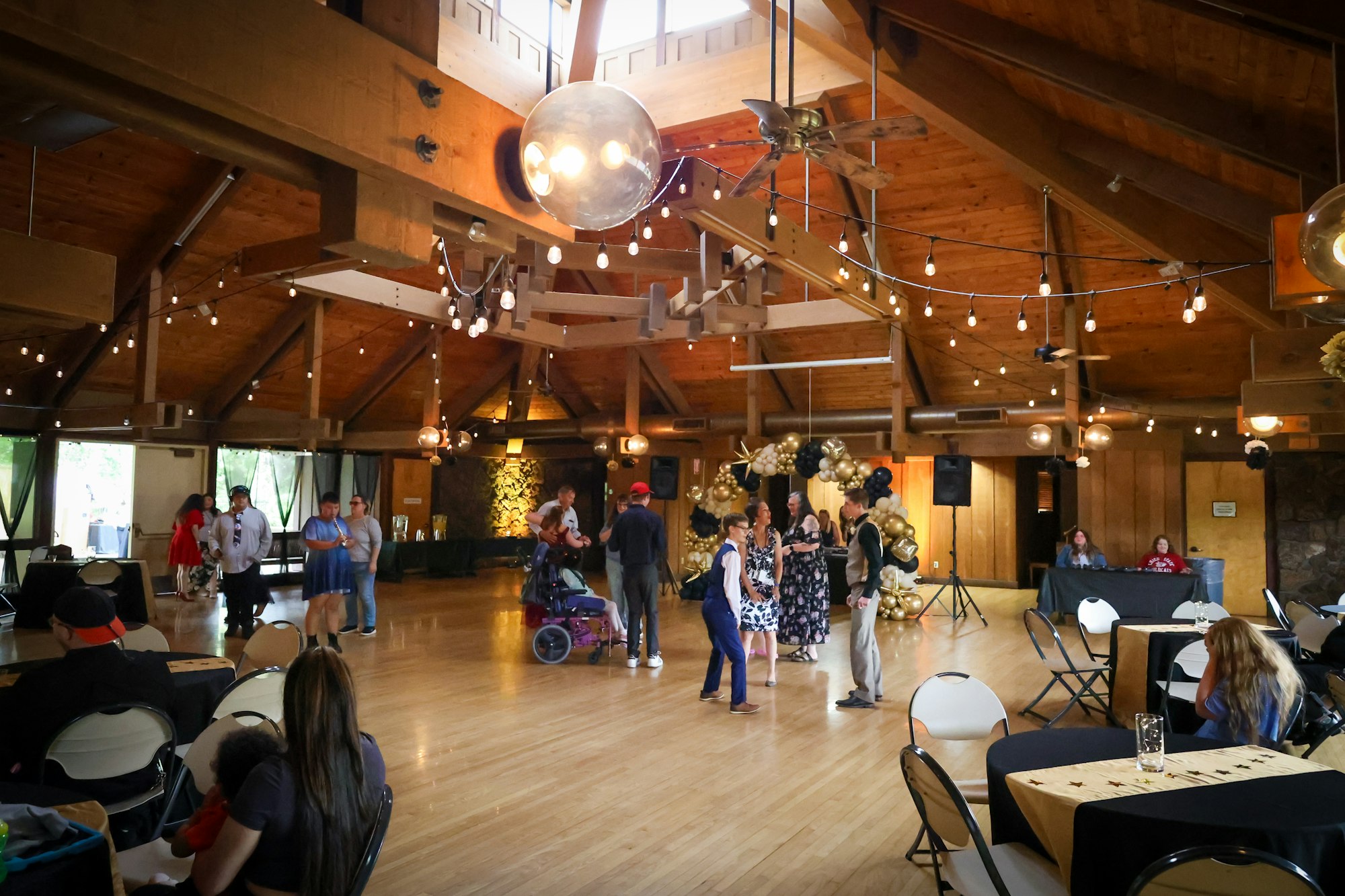 A spacious event hall with wooden architecture; people mingling, decorated tables, and festive lighting create a lively atmosphere.