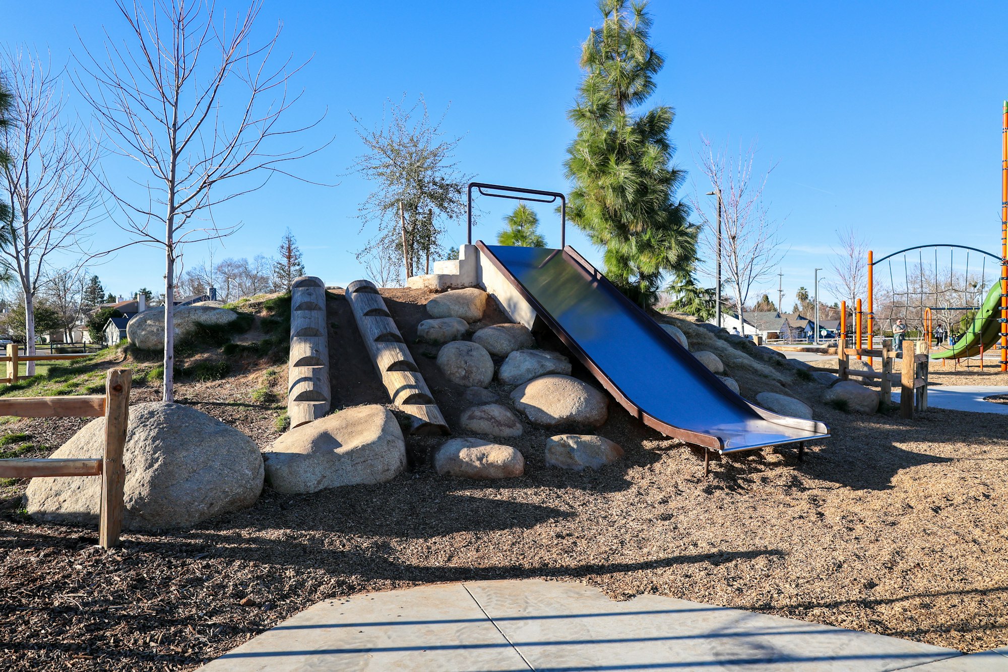 A park playground with a slide, climbing structures, rocks, and surrounding trees under a clear blue sky.