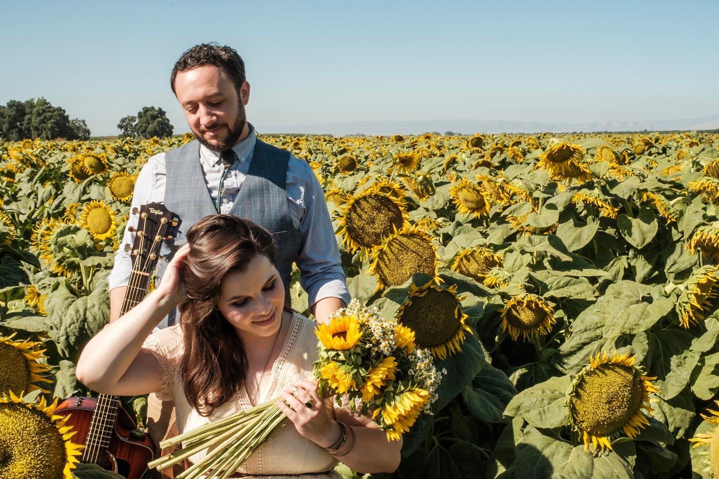 A couple in a sunflower field, holding a bouquet and guitar, enjoying a sunny day.