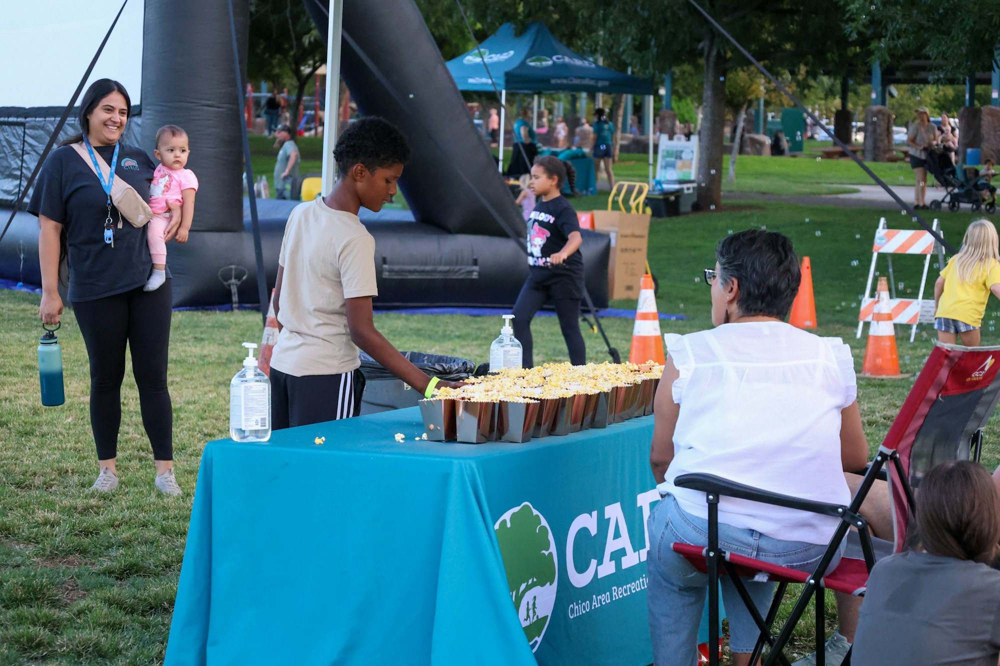 People at an outdoor event, a table with popcorn, a woman with a baby, and a kid in front of an inflatable structure.