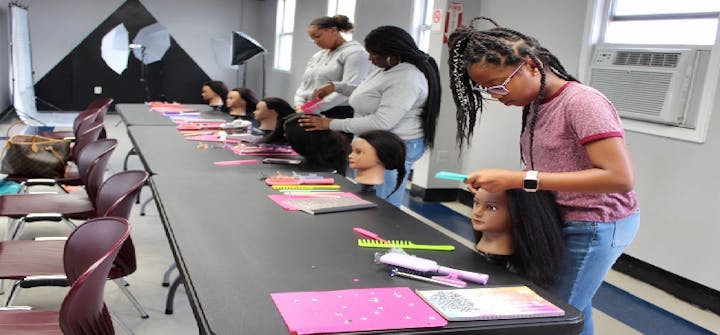 People practicing hairstyling on mannequin heads at a training table with combs and notebooks.