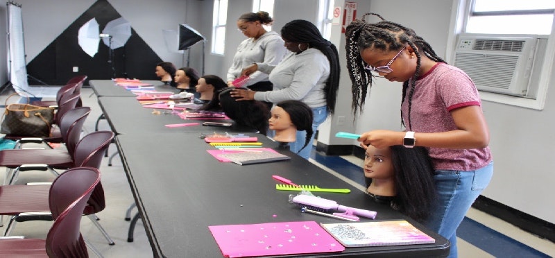 People practicing hairstyling on mannequin heads at a training table with combs and notebooks.