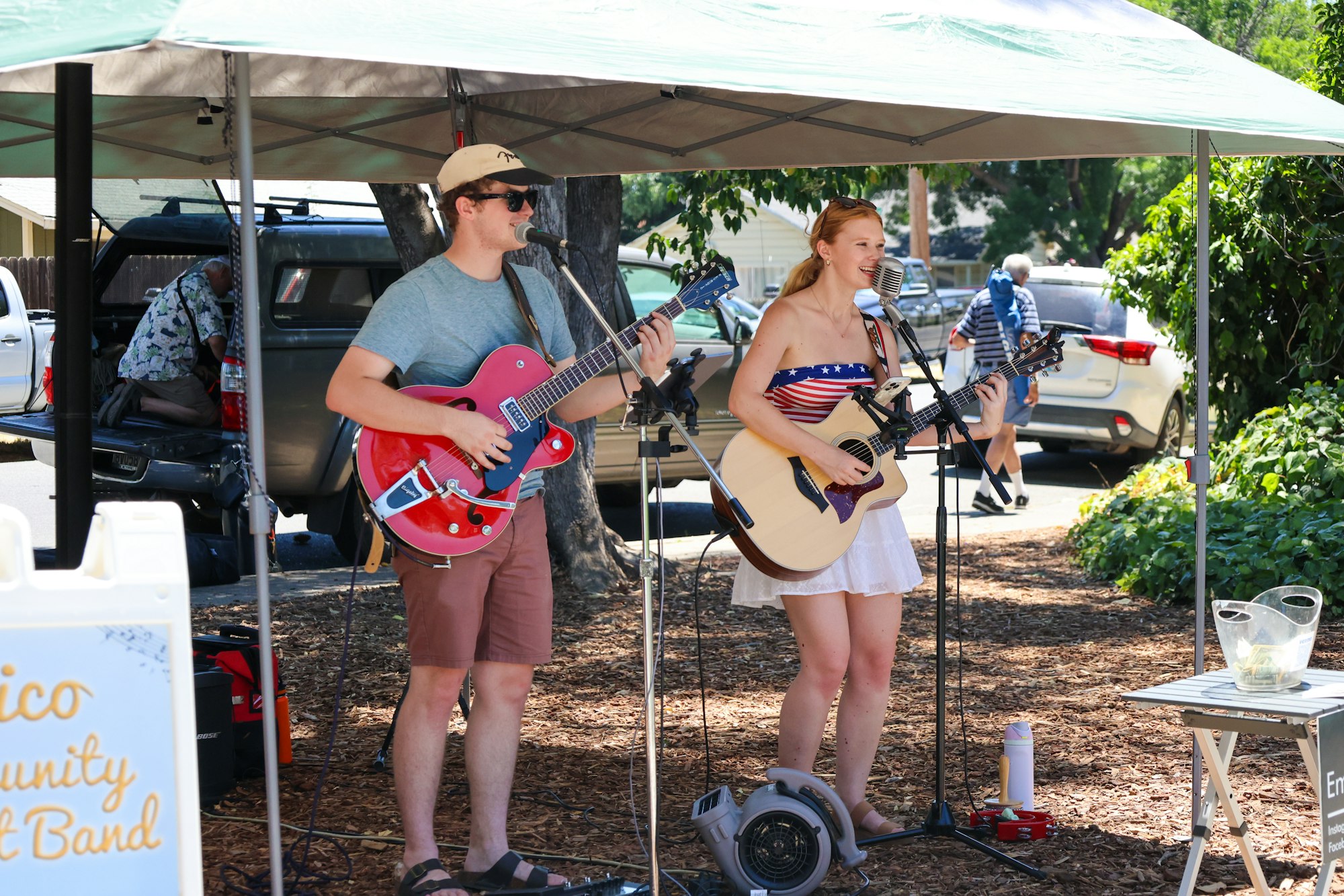 Two musicians performing outdoors under a canopy with guitars and microphones.