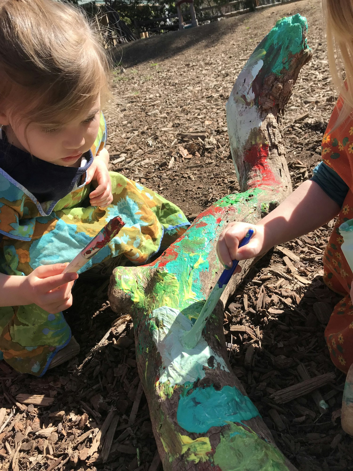 Children painting a large piece of wood outdoors with colorful paint.