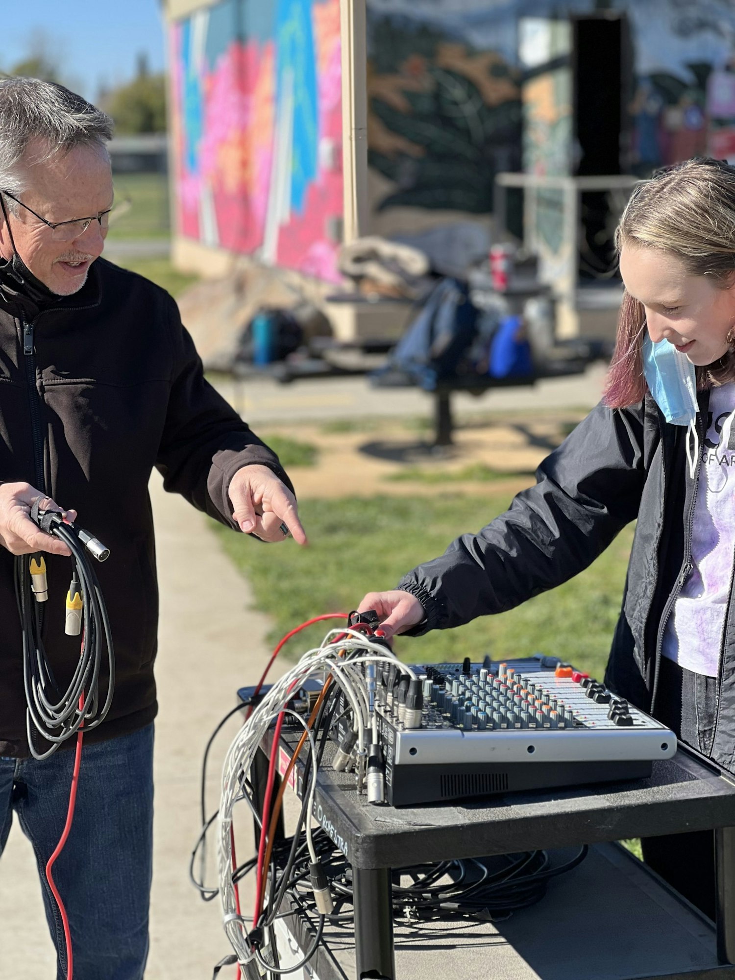 A man and a girl are working together on connecting audio equipment outdoors, with a vibrant mural in the background.