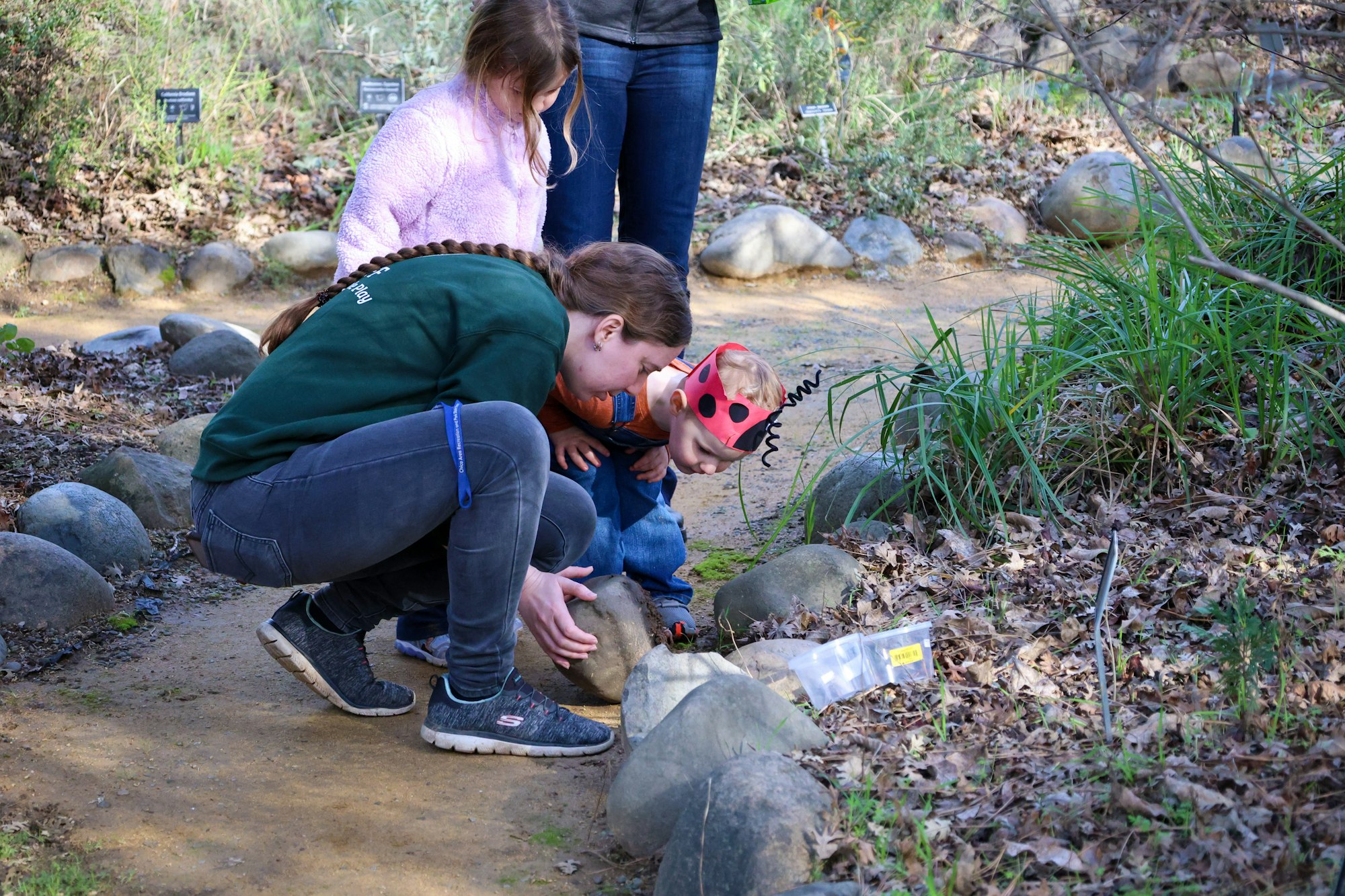 Children and an adult are observing an object on the ground in a natural setting, engaging in exploration and learning.