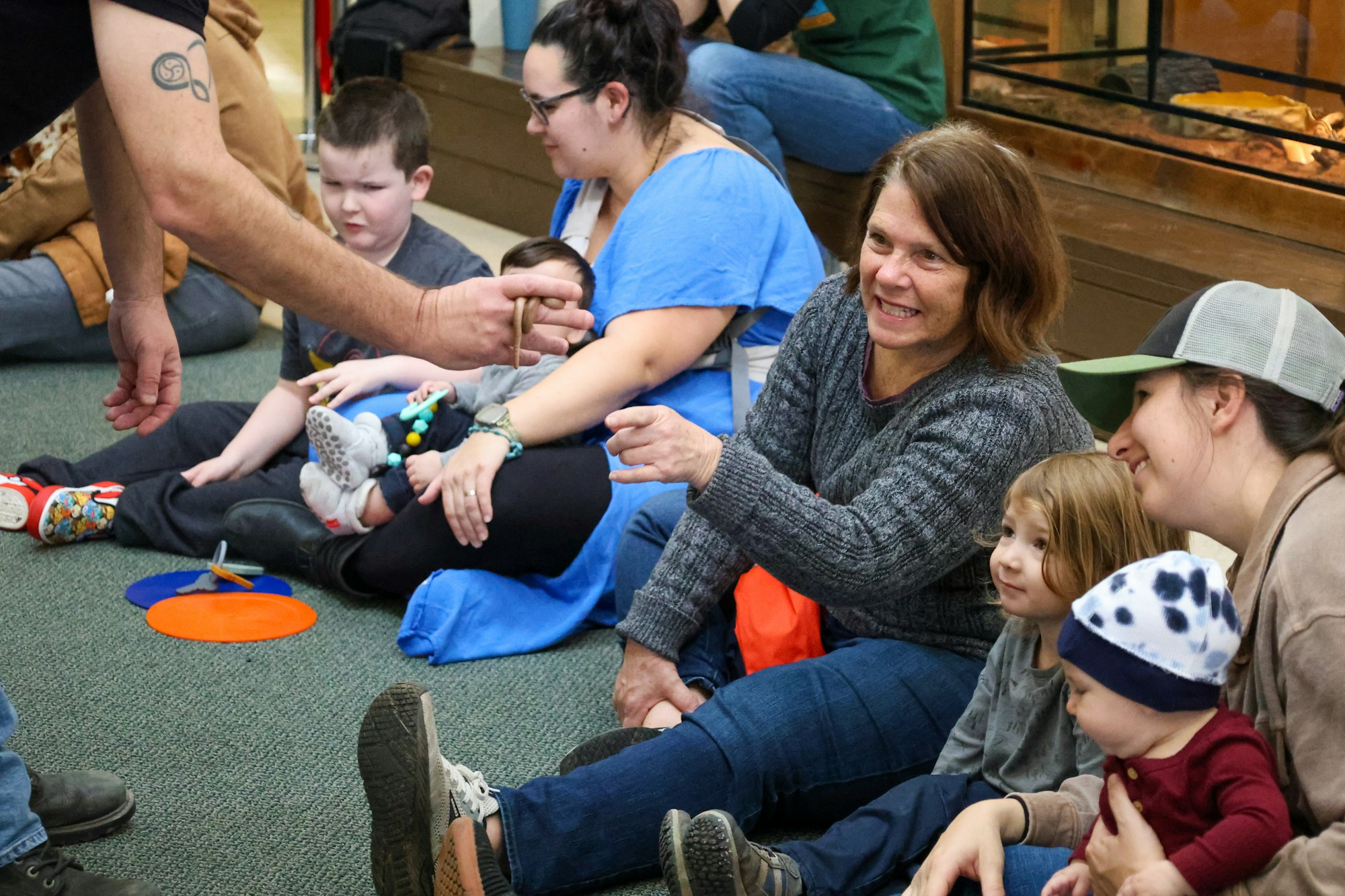 A group of adults and children are sitting on the floor, engaged and smiling, with someone holding a small object.
