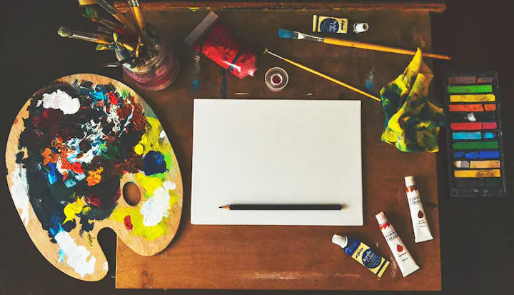 An artist's workspace featuring a paint palette, brushes, a blank canvas, and various paints and pastels.