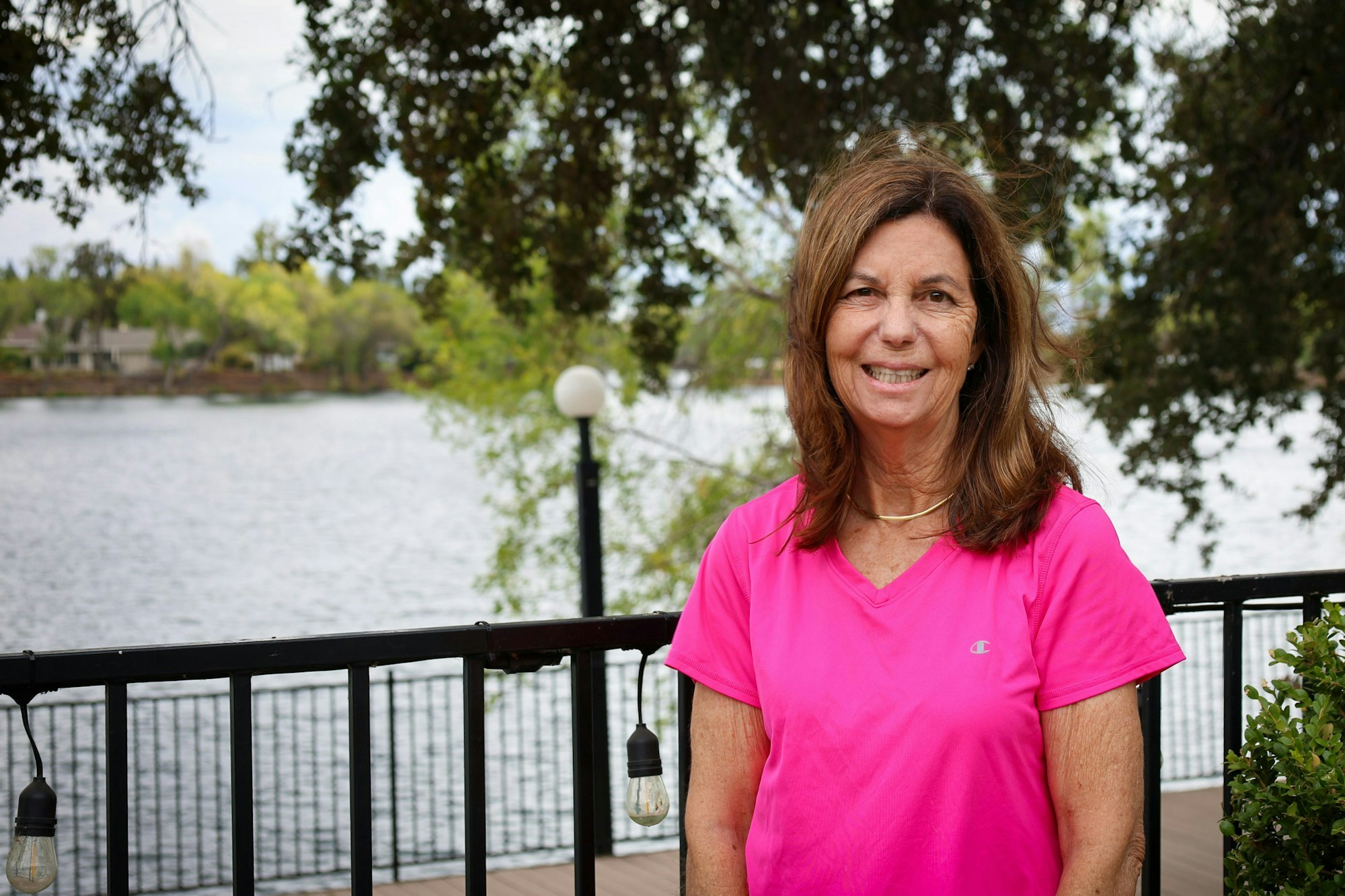A woman in a pink T-shirt stands by a lake, smiling, with trees and a railing in the background.