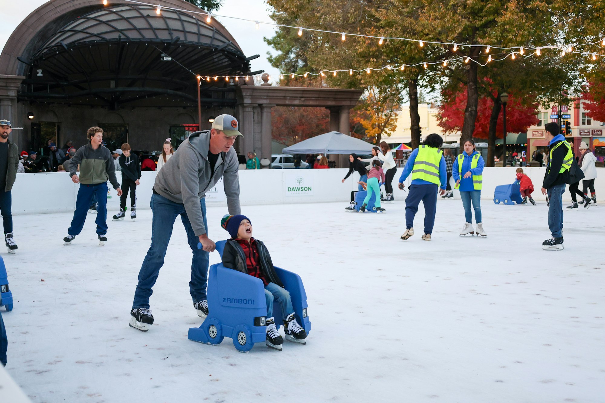 People ice skating on an outdoor rink; a man pushes a child in a blue sled.