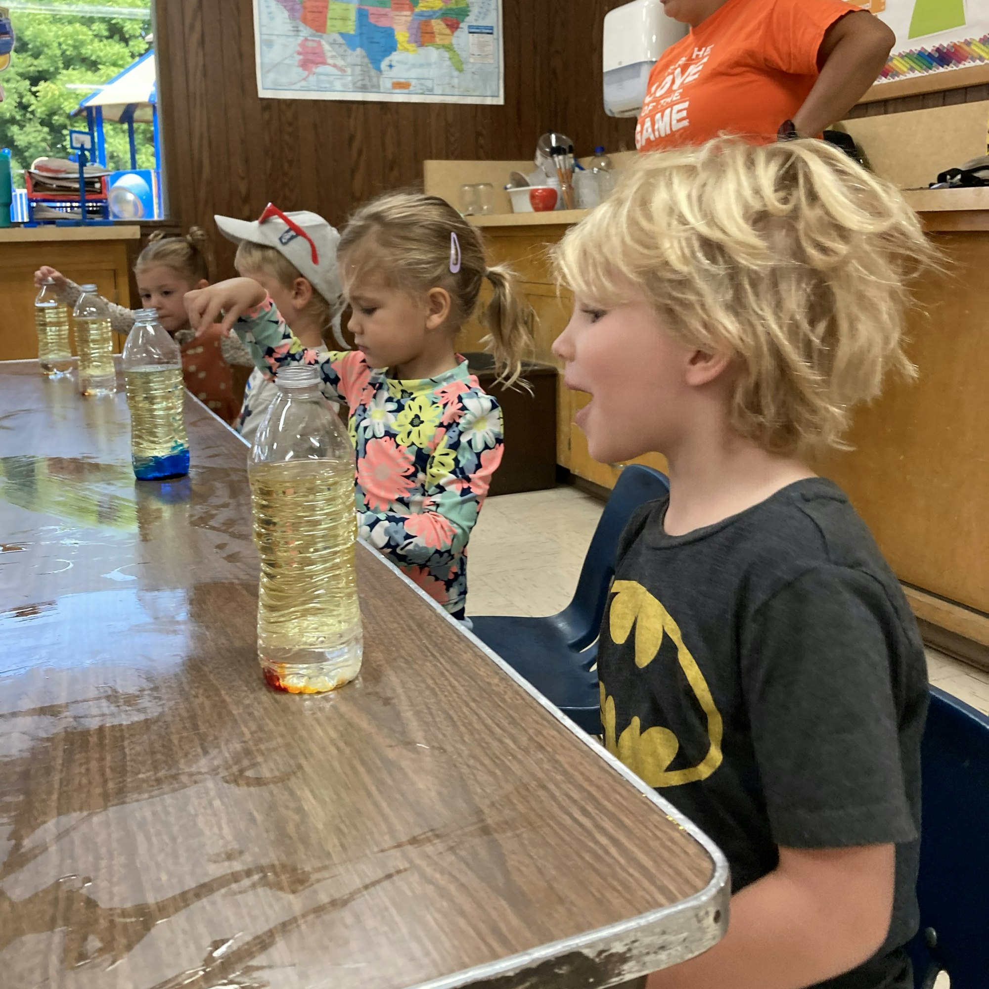 Children are engaged in a hands-on activity with bottles of liquid on a table, exploring colors and reactions.