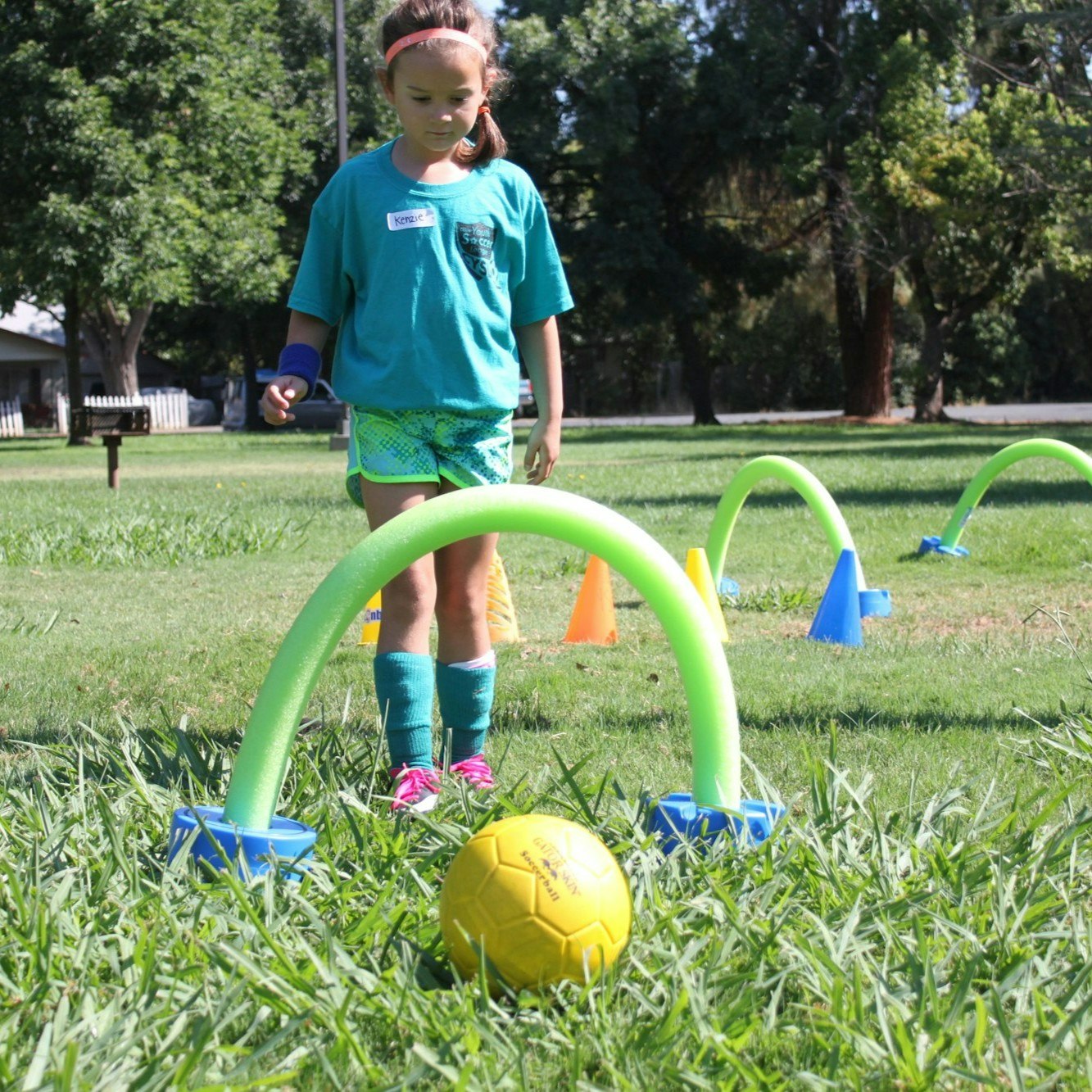 A girl in a teal shirt and shorts navigates a grassy area with colorful cones, foam arches, and a yellow soccer ball.
