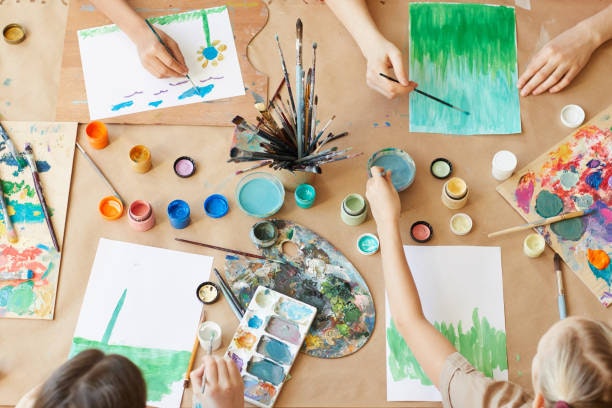 A creative workspace with hands painting on paper, brushes, and colorful paints arranged on a table.