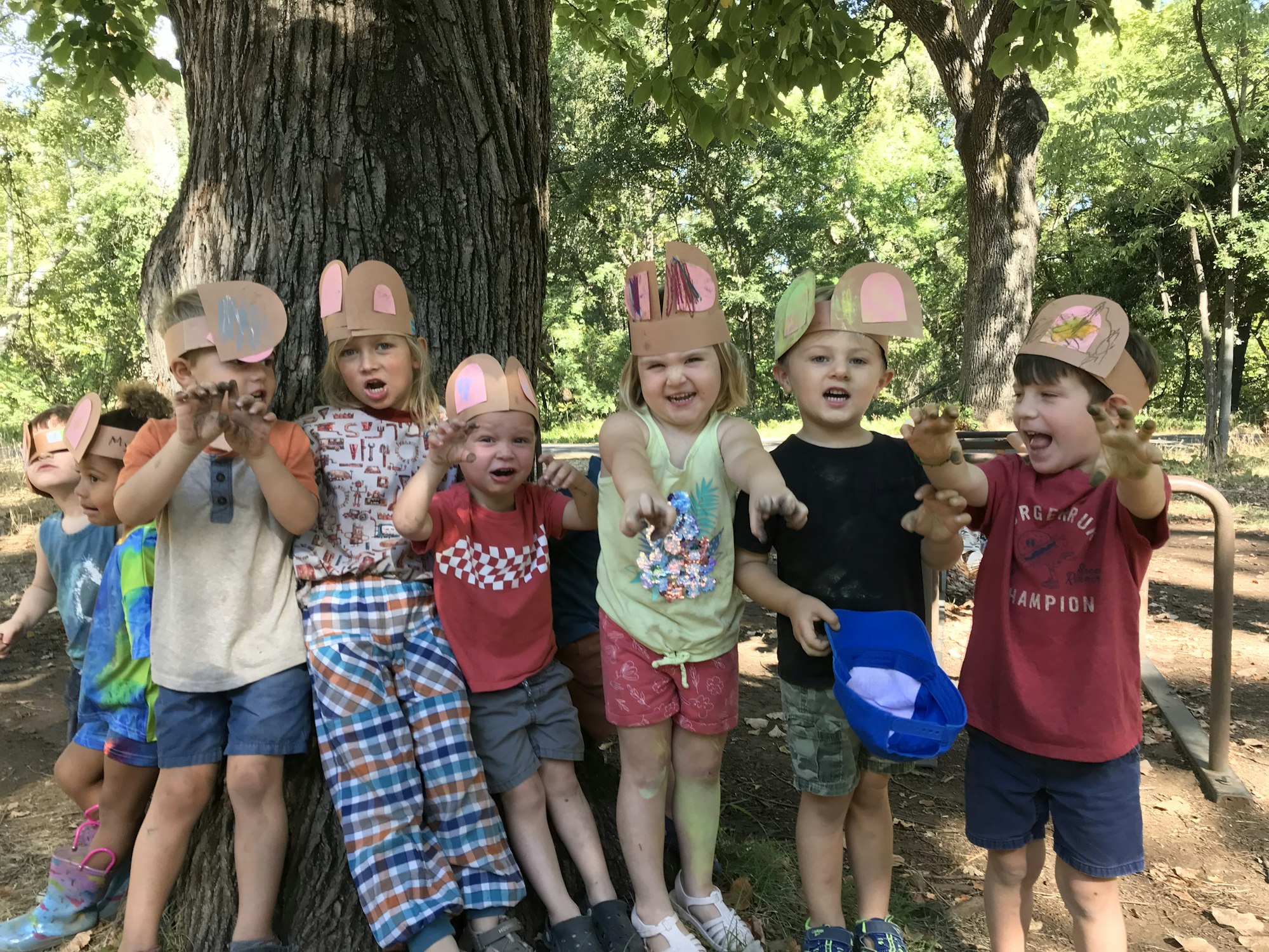 A group of children wearing handmade animal ear headbands, posing playfully under a tree.