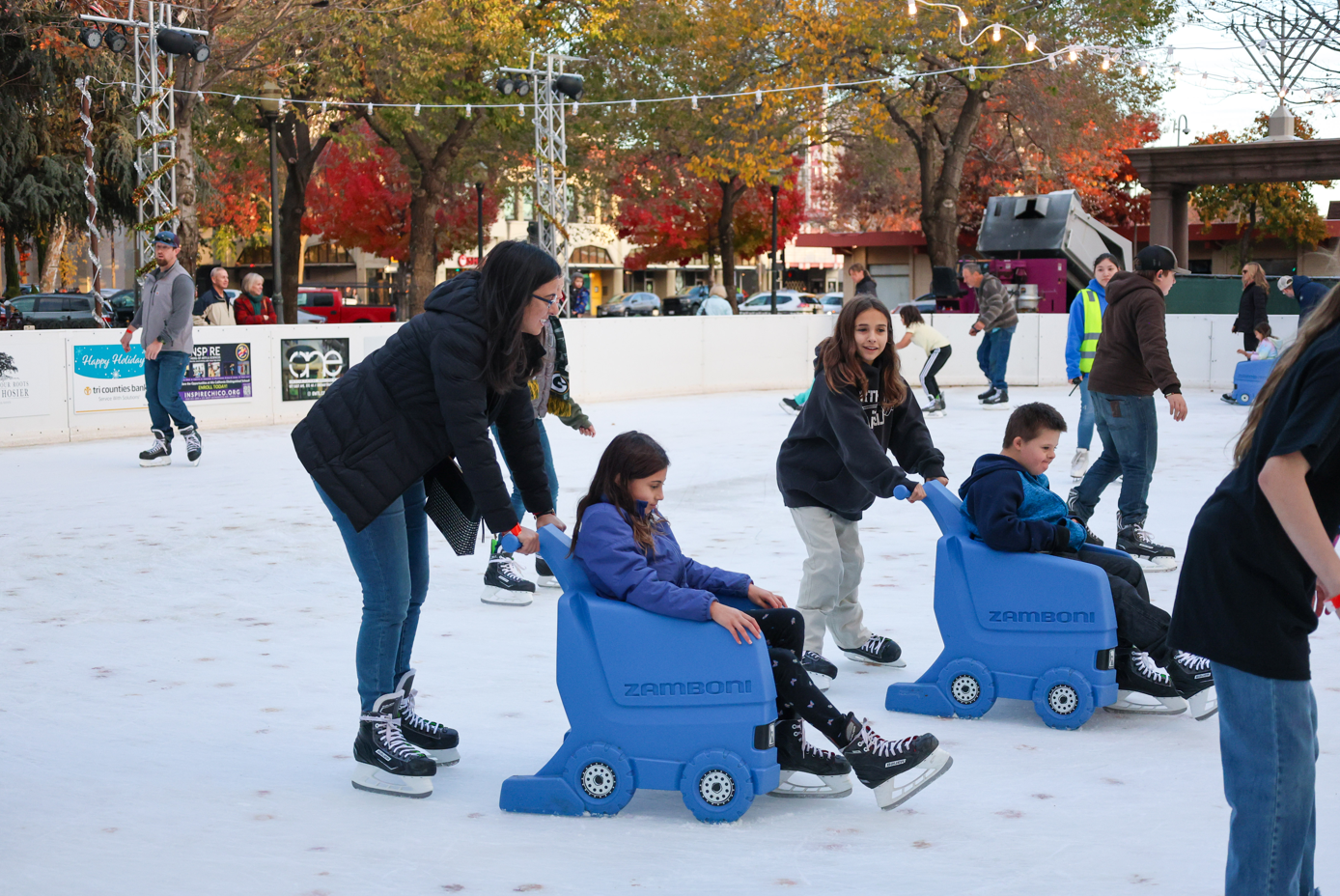 A group of people ice skating outdoors, some pushing children in blue Zamboni-themed skating aids. Fall colors in the background.