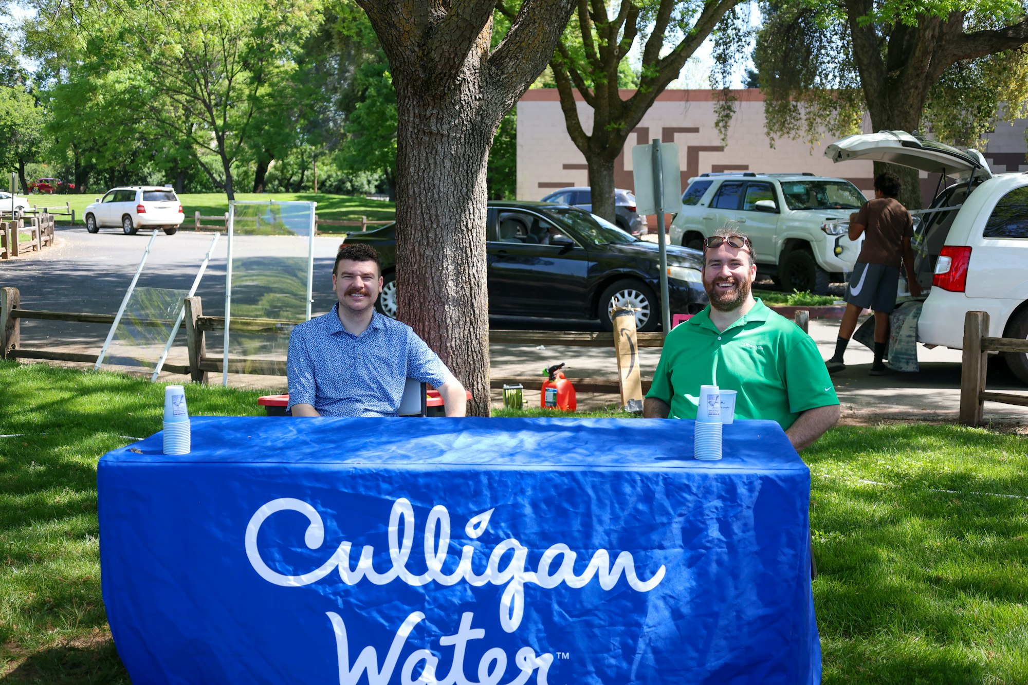 Two people sitting at a table with a "Culligan Water" cloth outdoors, with cars and trees in the background.