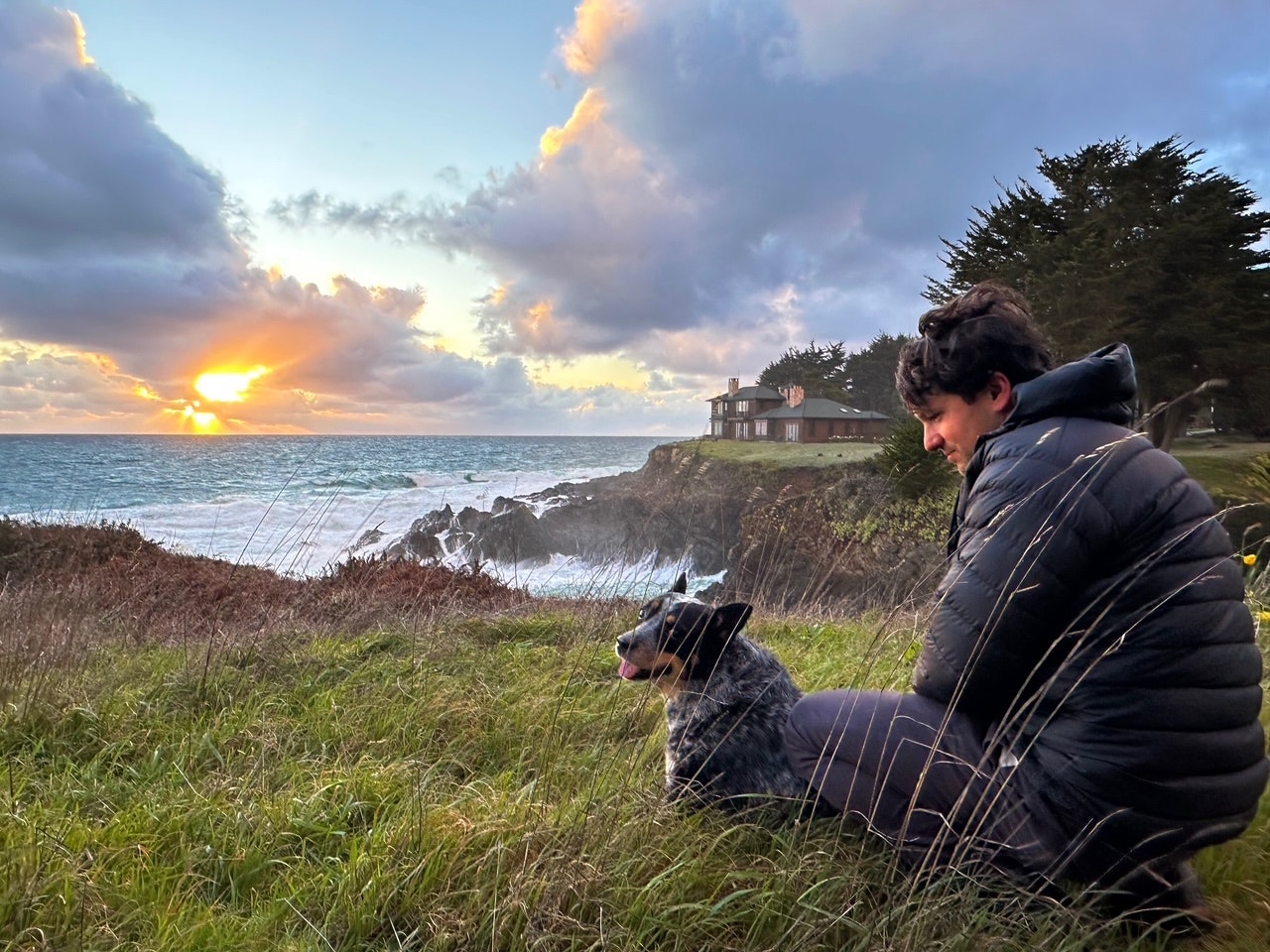 A person and a dog sit on grass by the ocean, watching a vibrant sunset with clouds and a house in the background.