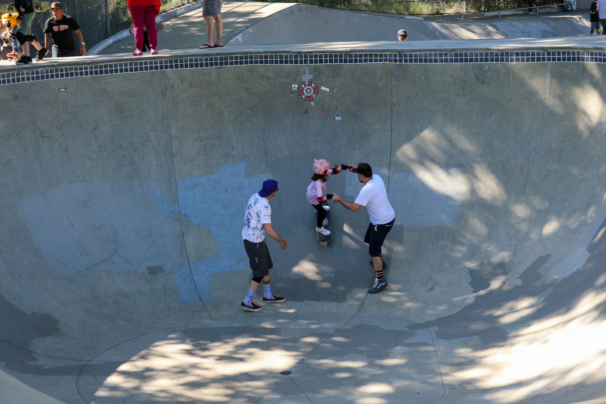 Skatepark with people, child learning to skate, adults standing and watching.