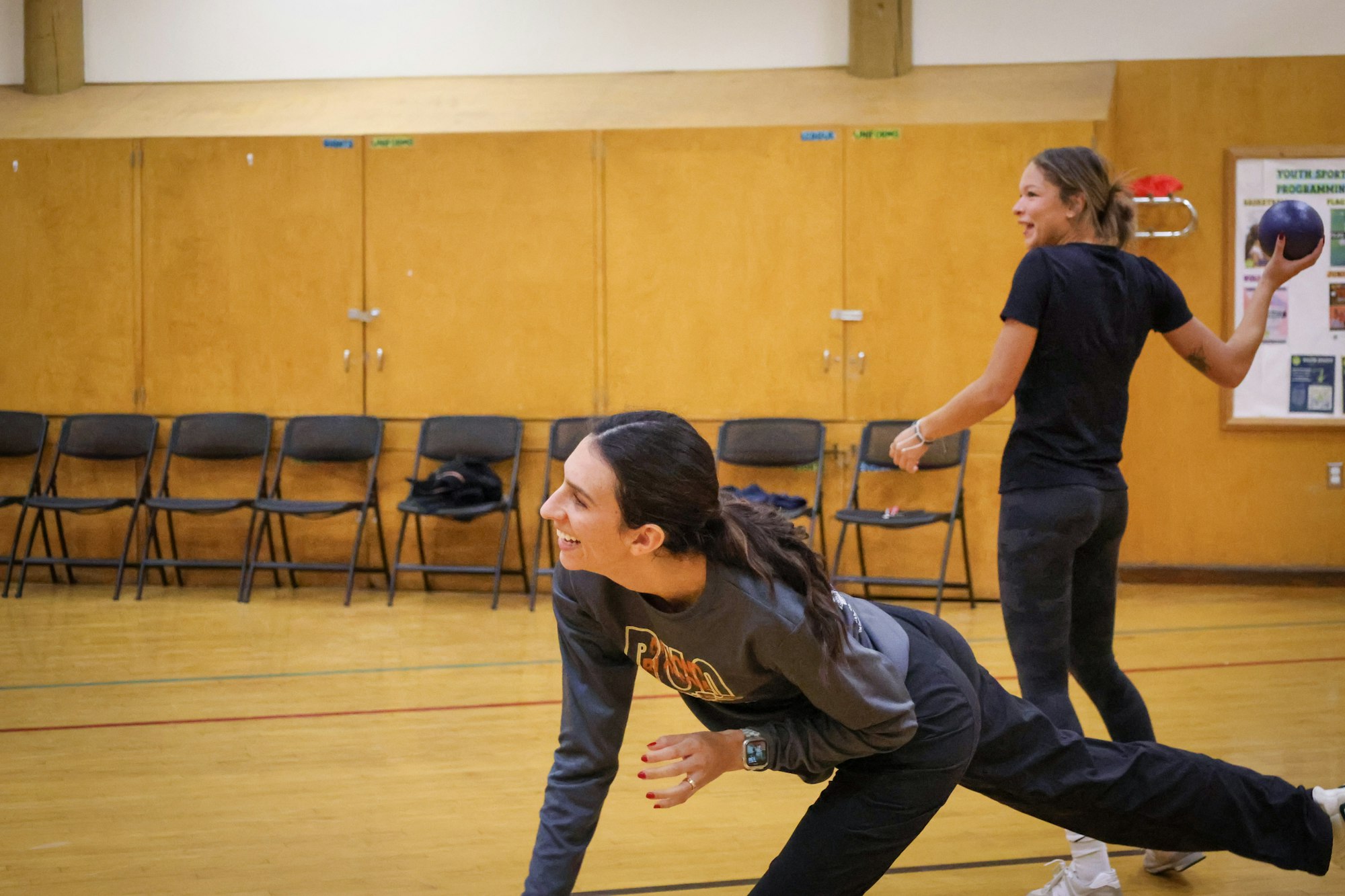 Two women are playing a game in a gym, one laughing while bending down, and the other preparing to throw a ball.