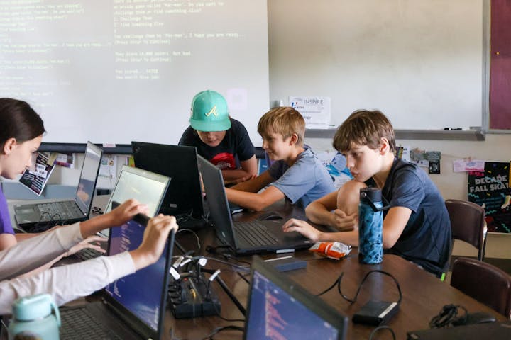 Kids are coding on laptops around a table in a classroom setting, with code projected on a screen.