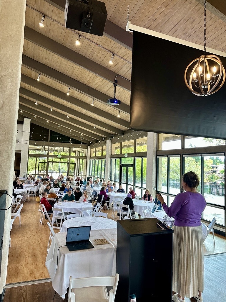 A speaker addresses an audience in a bright, spacious venue with white tables and large windows.